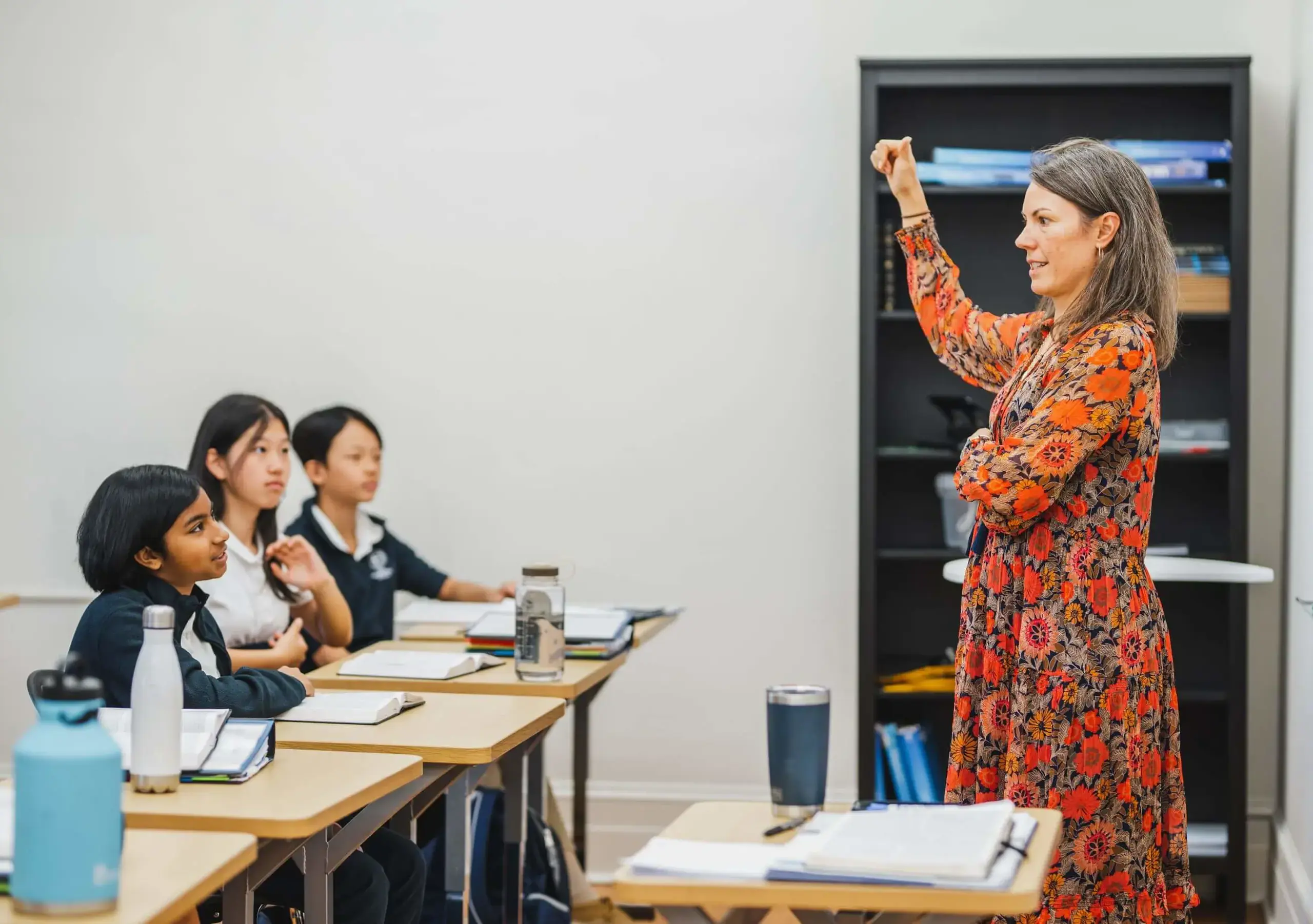 A teacher in a floral dress leads a class of students in a lesson.
